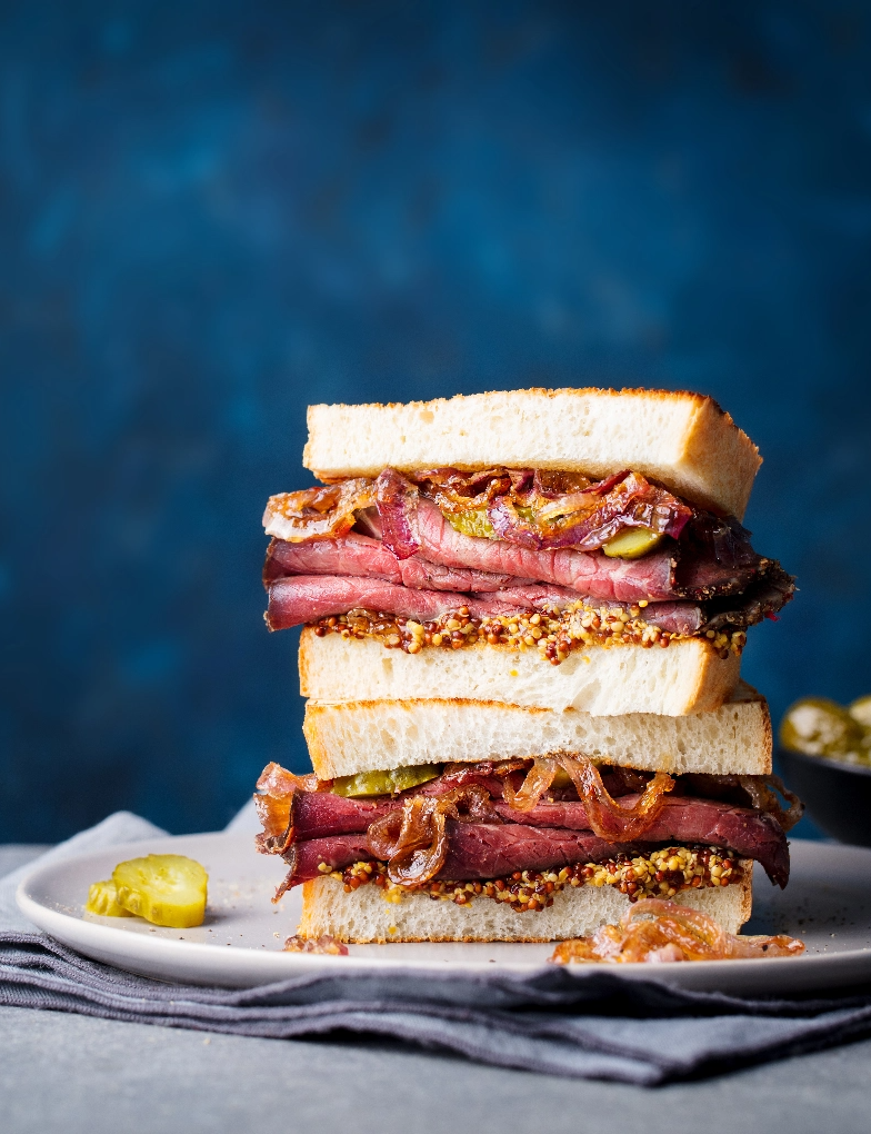 A roast beef sandwich stacked high with meat and grainy mustard, with pickles on the side