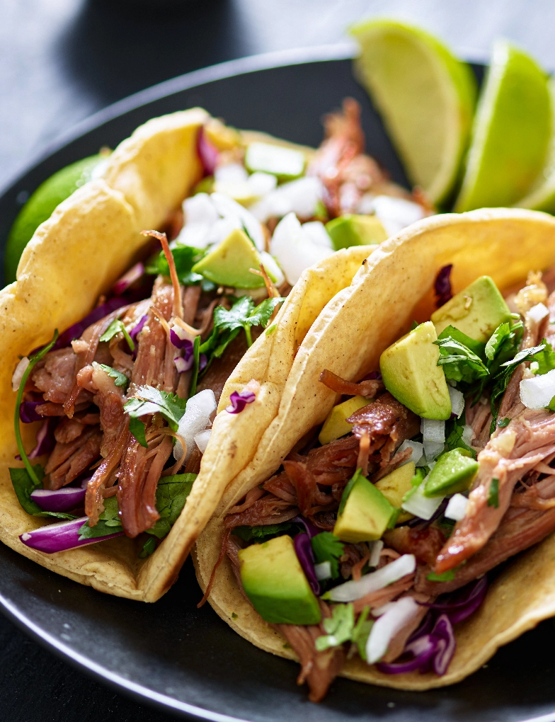Pulled pork tacos with fresh veggies and cilantro, served on a dark plate with lime wedges.