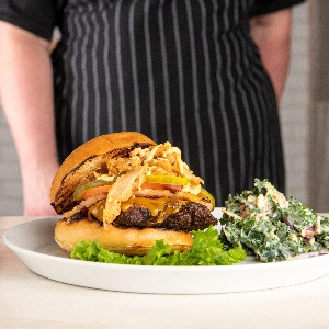 A chef stands behind a table with a plated burger and cole slaw.