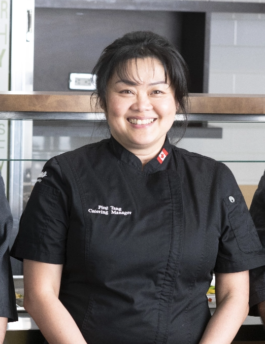 A female catering manager in black kitchen uniform smiles at the camera from a kitchen