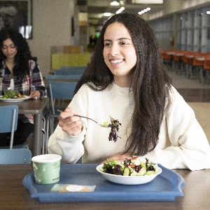 A young woman sits in a cafeteria eating from a bowl of salad on a blue tray.