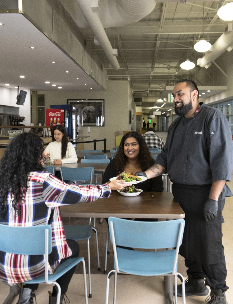 A Dana employee in kitchen uniform hands a bowl of salad to a female student sitting at a cafeteria table with another female student.