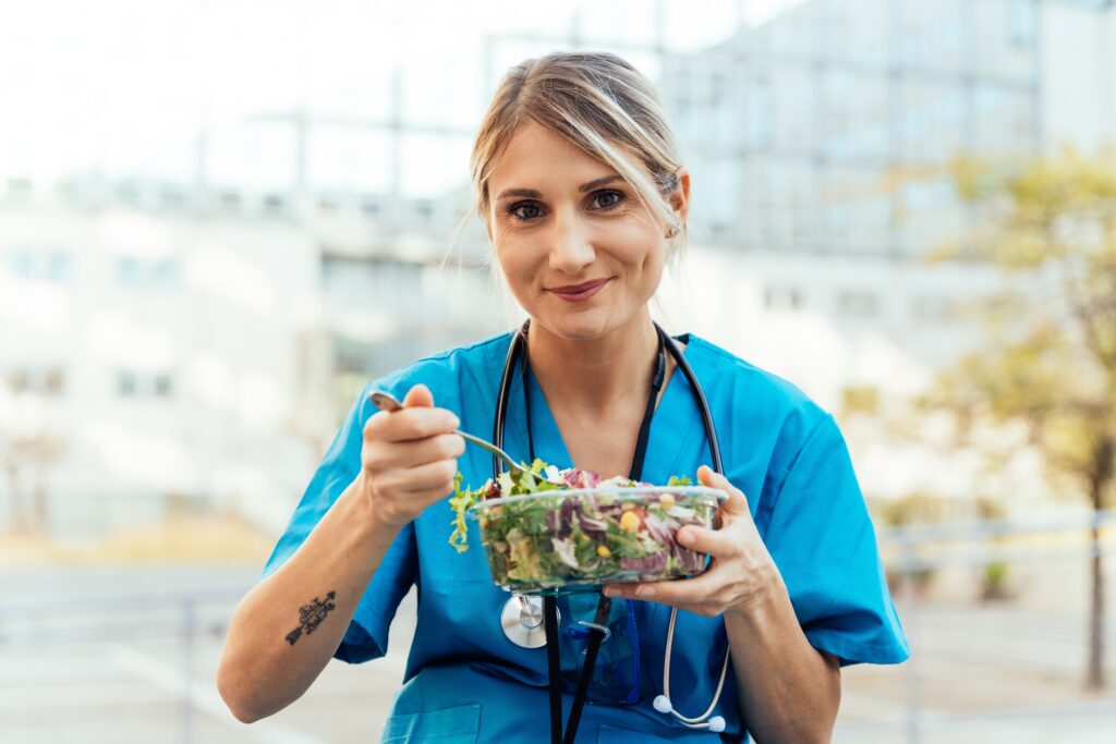 A woman in medical scrubs with a stethoscope around her neck eats a healthy-looking salad while outside.