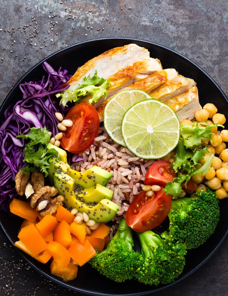 A brown rice bowl with a rainbow of veggies, meat and nuts sits on a dark stone countertop.